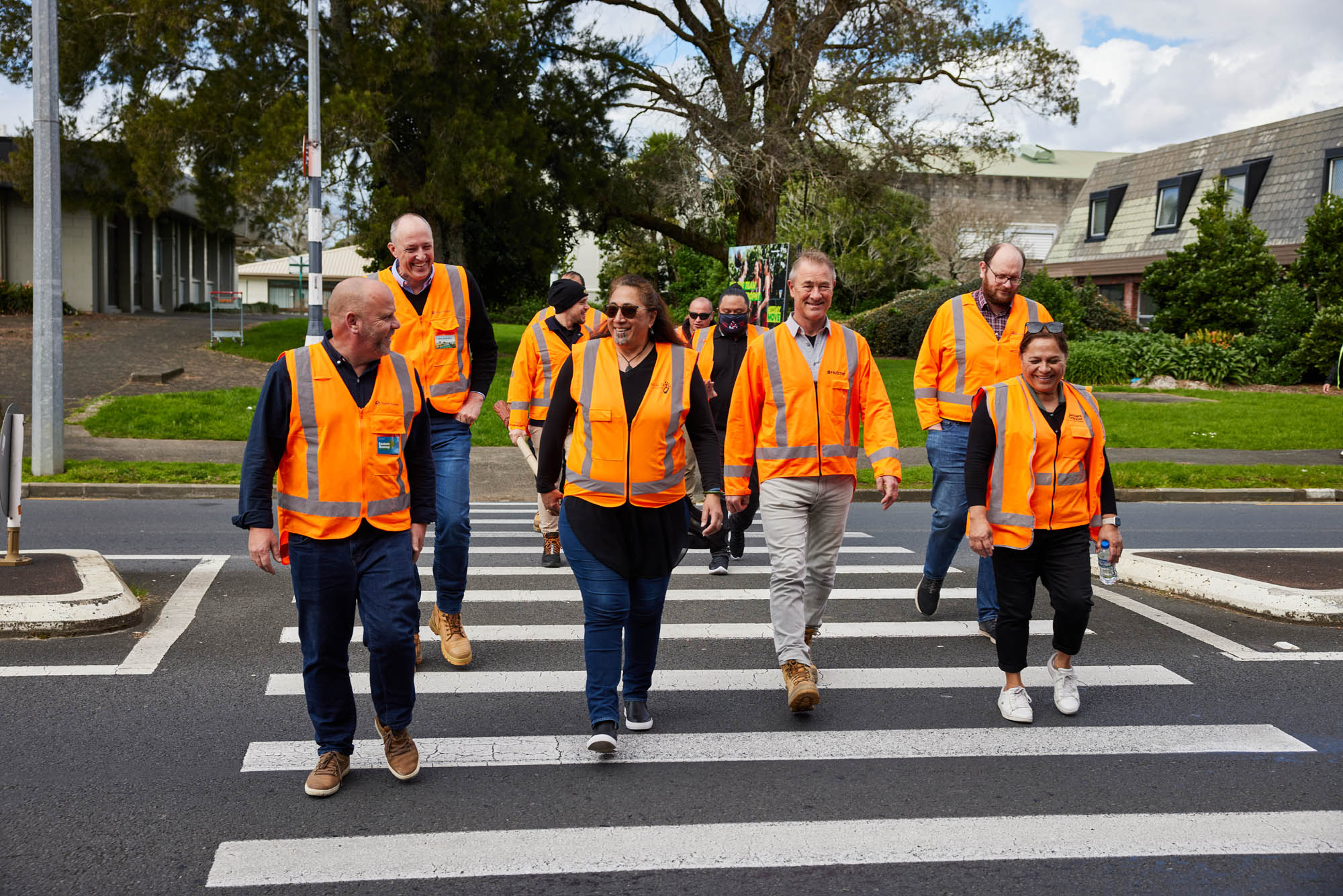 A group of people from the project team with mana whenua representatives wearing orange high vis vests crossing Reeves Road using a pedestrian crossing. A group of people from the project team with mana whenua representatives wearing orange high vis vests crossing Reeves Road using a pedestrian crossing.